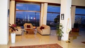 A spacious living room with a large window, a beige sofa, a coffee table, and a potted plant. - Taybeh Golden Hotel (Taybeh)