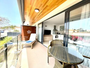Un balcón con una silla de mimbre y una mesa, una puerta corredera de vidrio que da a una cocina, y vistas al exterior.