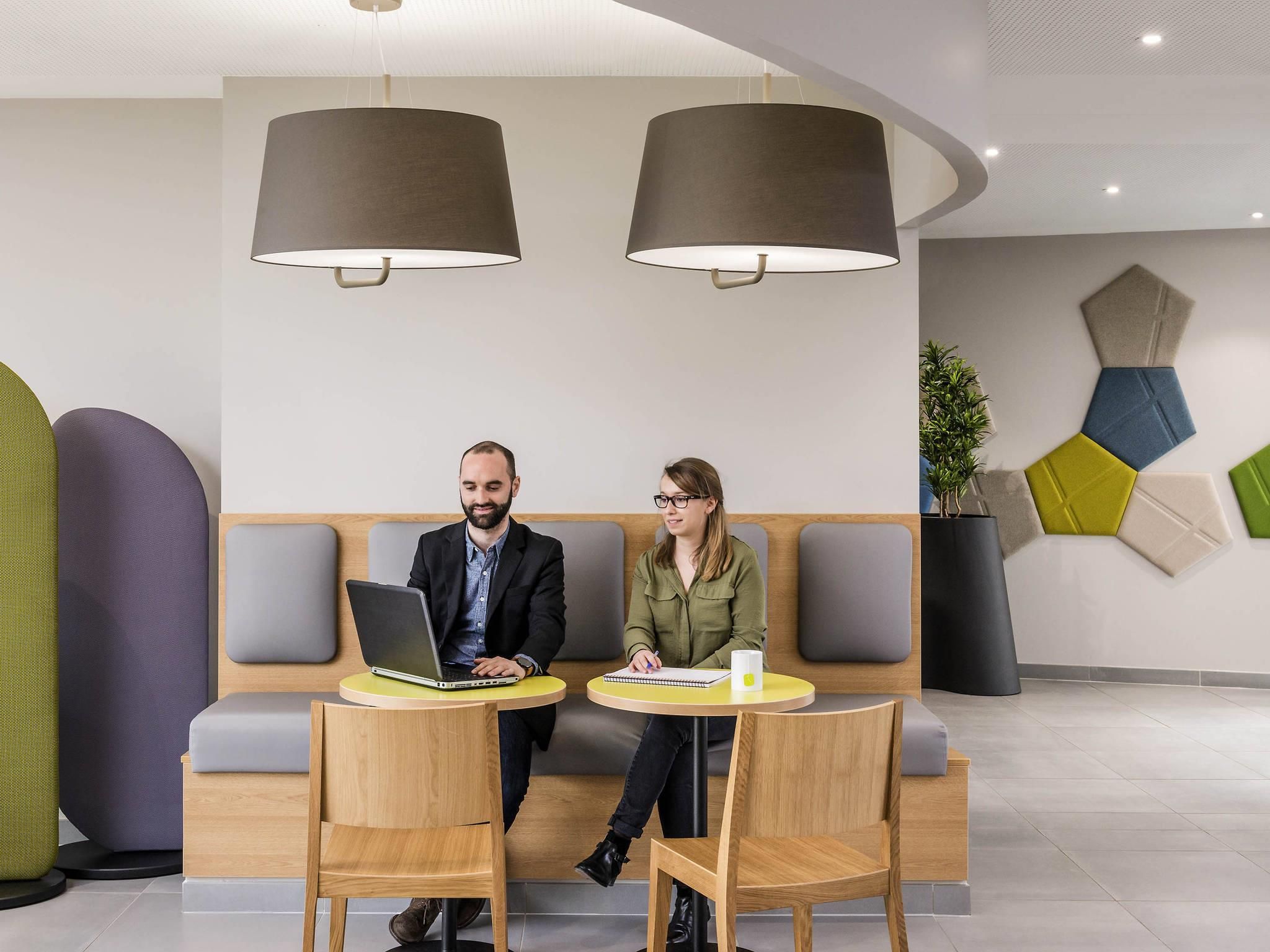 Deux personnes sont assises à une table, devant des ordinateurs portables, dans un intérieur moderne agrémenté d’un canapé et de luminaires suspendus.