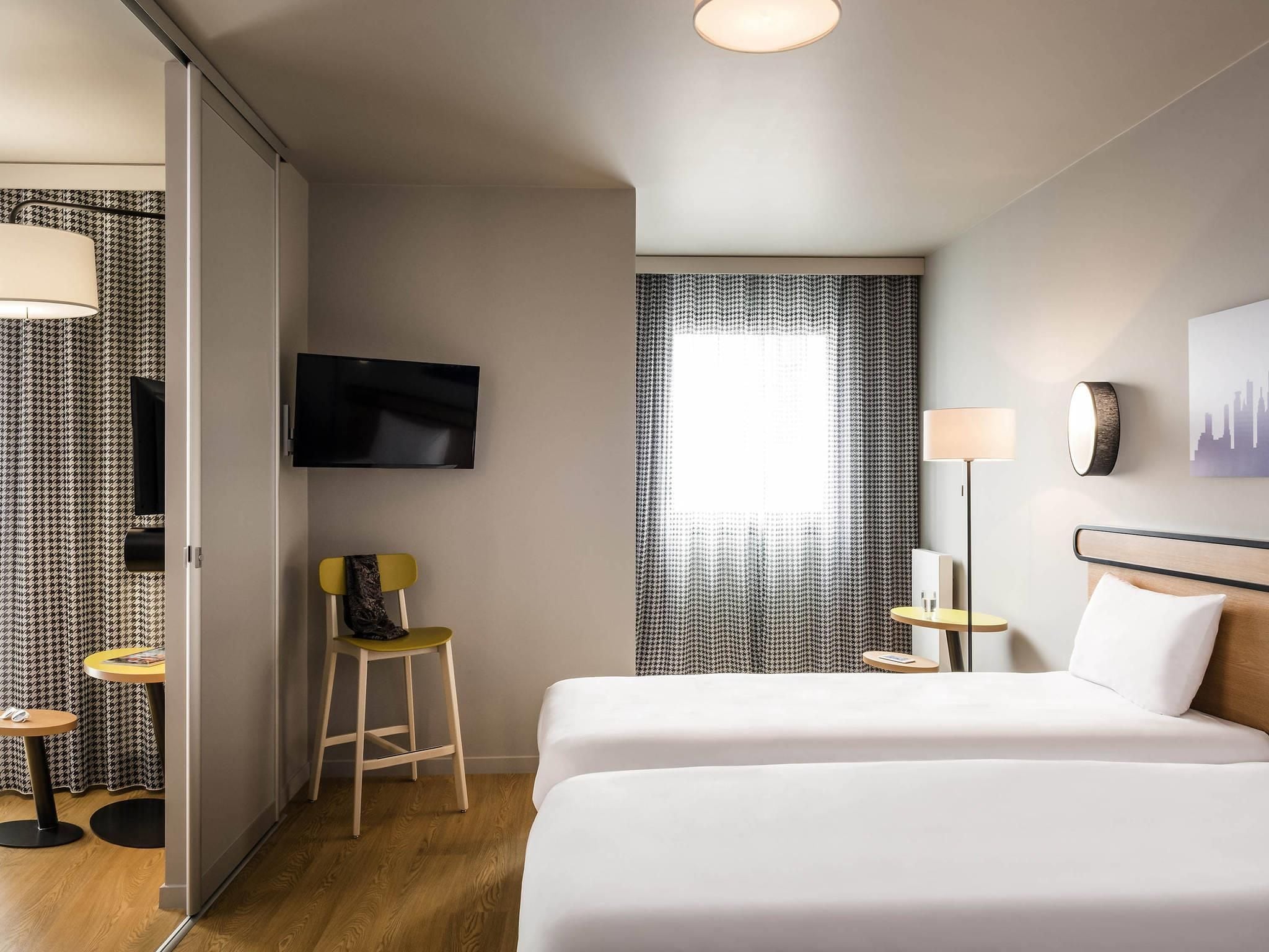Une chambre d’hôtel avec un lit, une télévision, une chaise, une table et une fenêtre avec des rideaux.