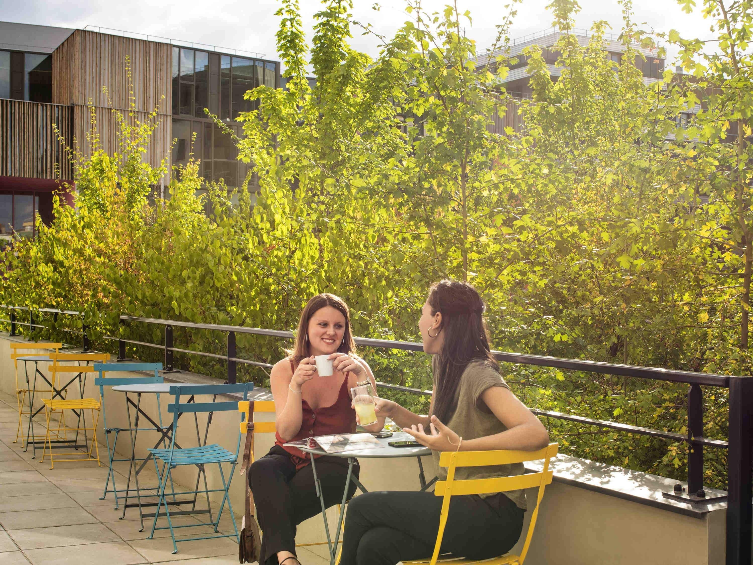 Deux personnes sont assises à une table à l’extérieur, avec un bâtiment et des arbres en arrière-plan.