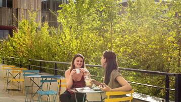 Deux personnes sont assises à une table à l’extérieur, avec un bâtiment et des arbres en arrière-plan.