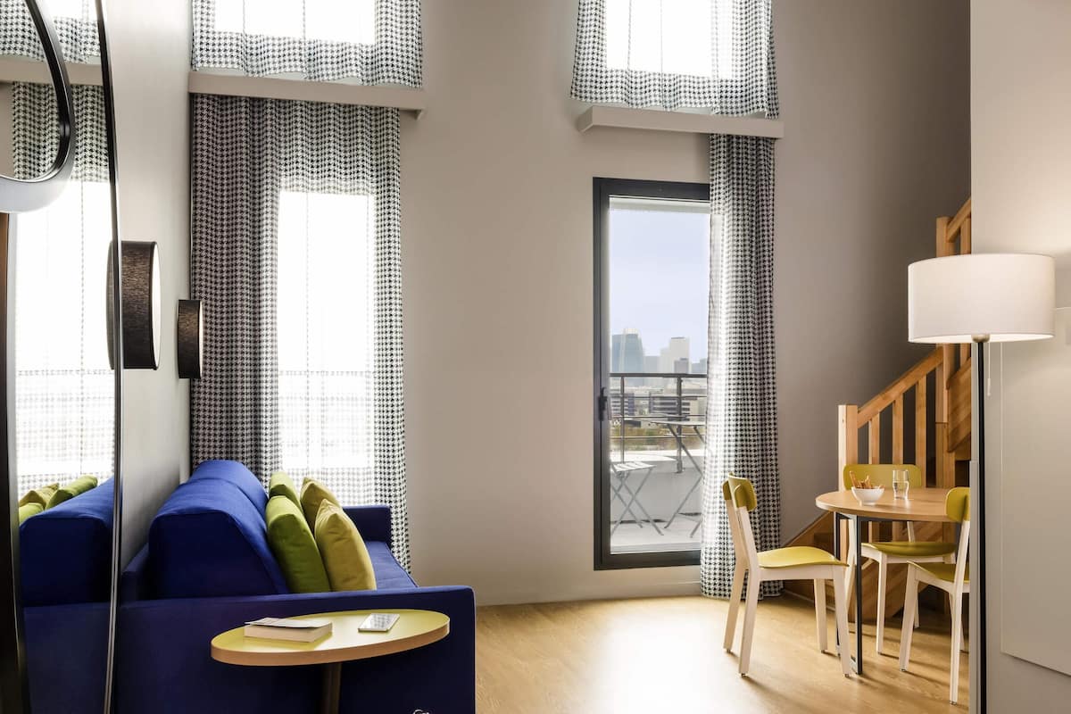 Une pièce moderne avec un canapé bleu, une petite table ronde, une chaise, une lampe et une vue sur la ville par une fenêtre.