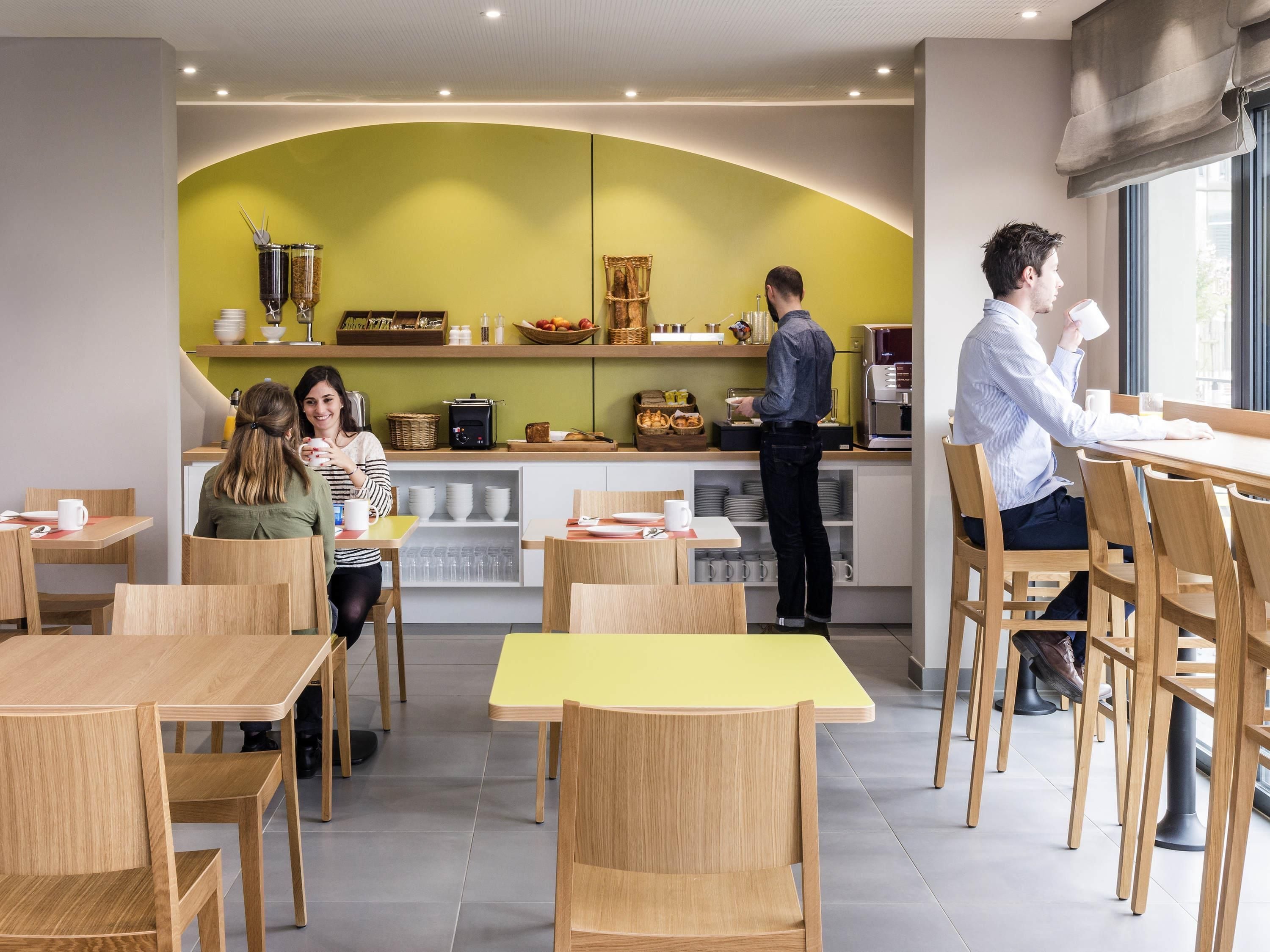 A modern cafe with wooden tables and chairs, a green accent wall, and a counter with food and drink items.