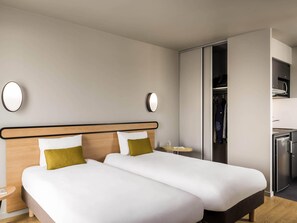 A hotel room with two beds, a wardrobe, a small table, and a built-in fridge.