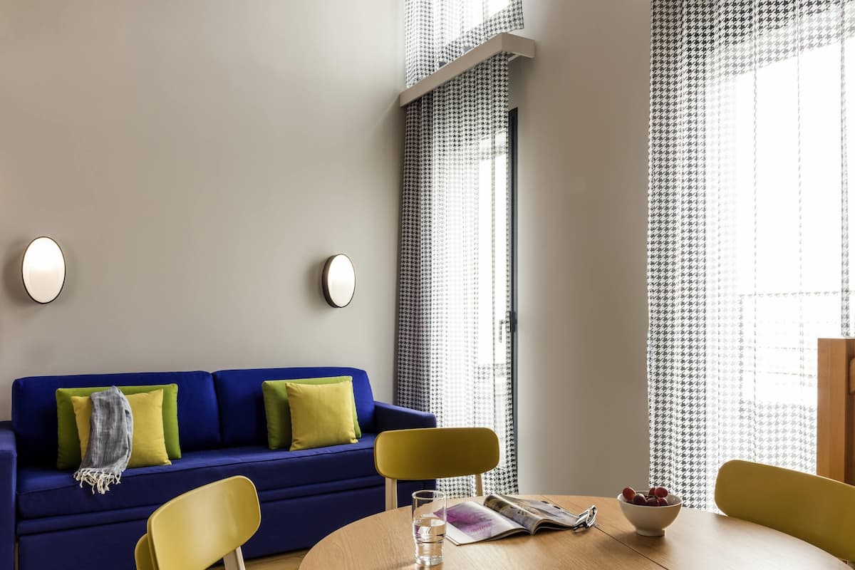 A modern living room with a blue sofa, yellow chairs, a wooden table, and a window with patterned curtains.