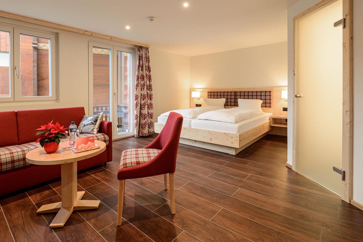 Habitación de hotel con sofá rojo, una mesa redonda de madera con una planta, una silla roja, cama con ropa blanca y suelo de madera.