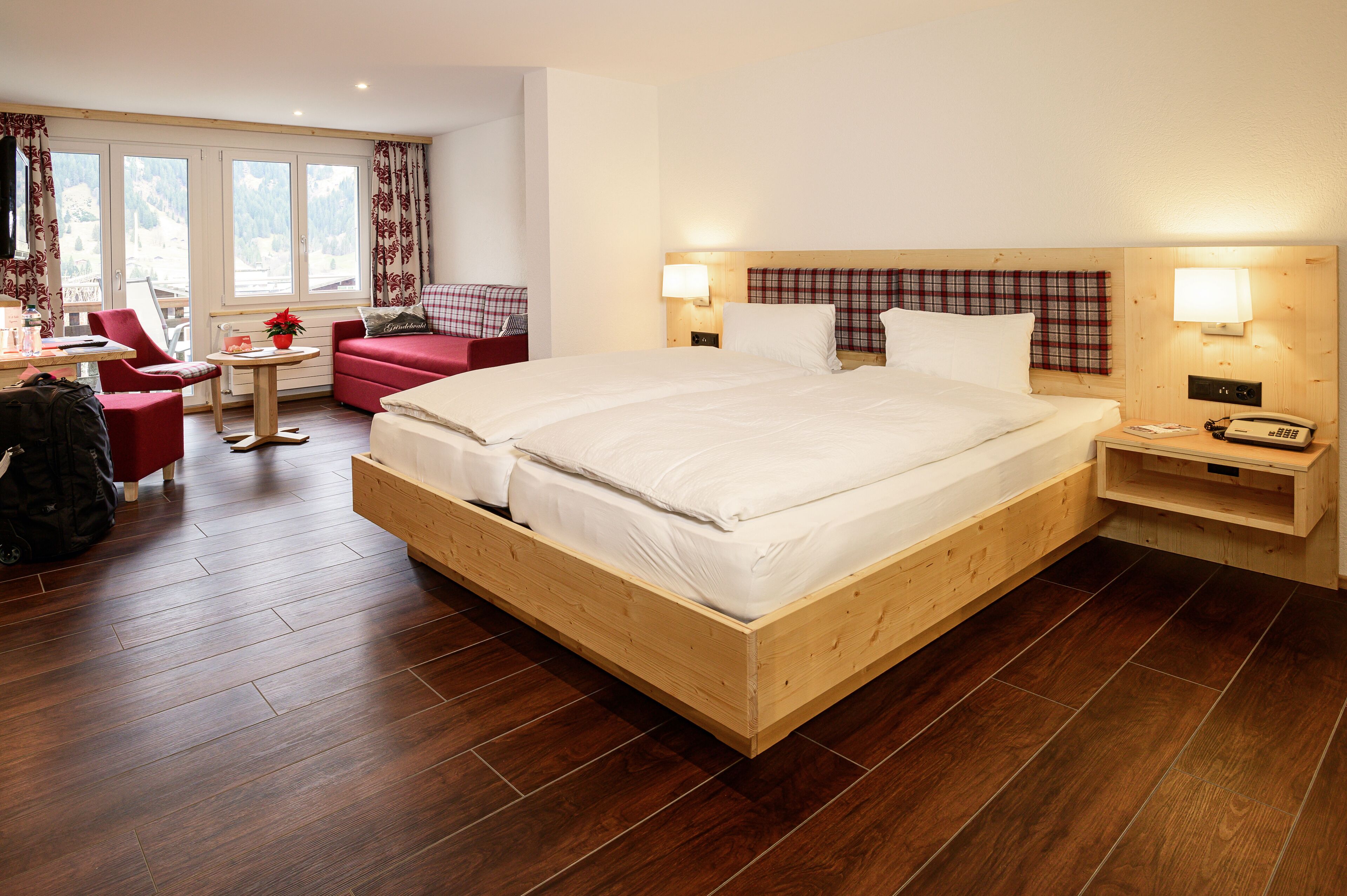 Una cama de madera con ropa blanca, un sillón rojo, una mesita con una planta y vistas a montañas a través de amplios ventanales.