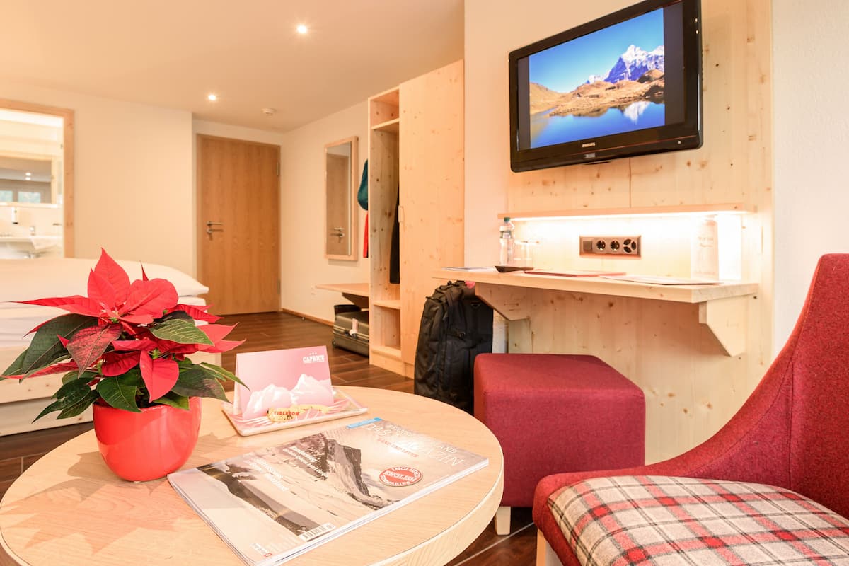 Una habitación de hotel moderna con interior de madera, un televisor de pantalla plana montado en la pared, una mesa redonda con una revista y una planta en maceta roja, y un sillón rojo.