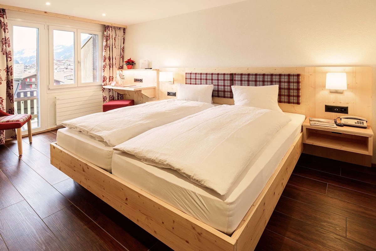 Una cama doble con cabecera de madera, un escritorio con silla, una mesita de noche con lámpara, y vistas a la montaña a través de la ventana.