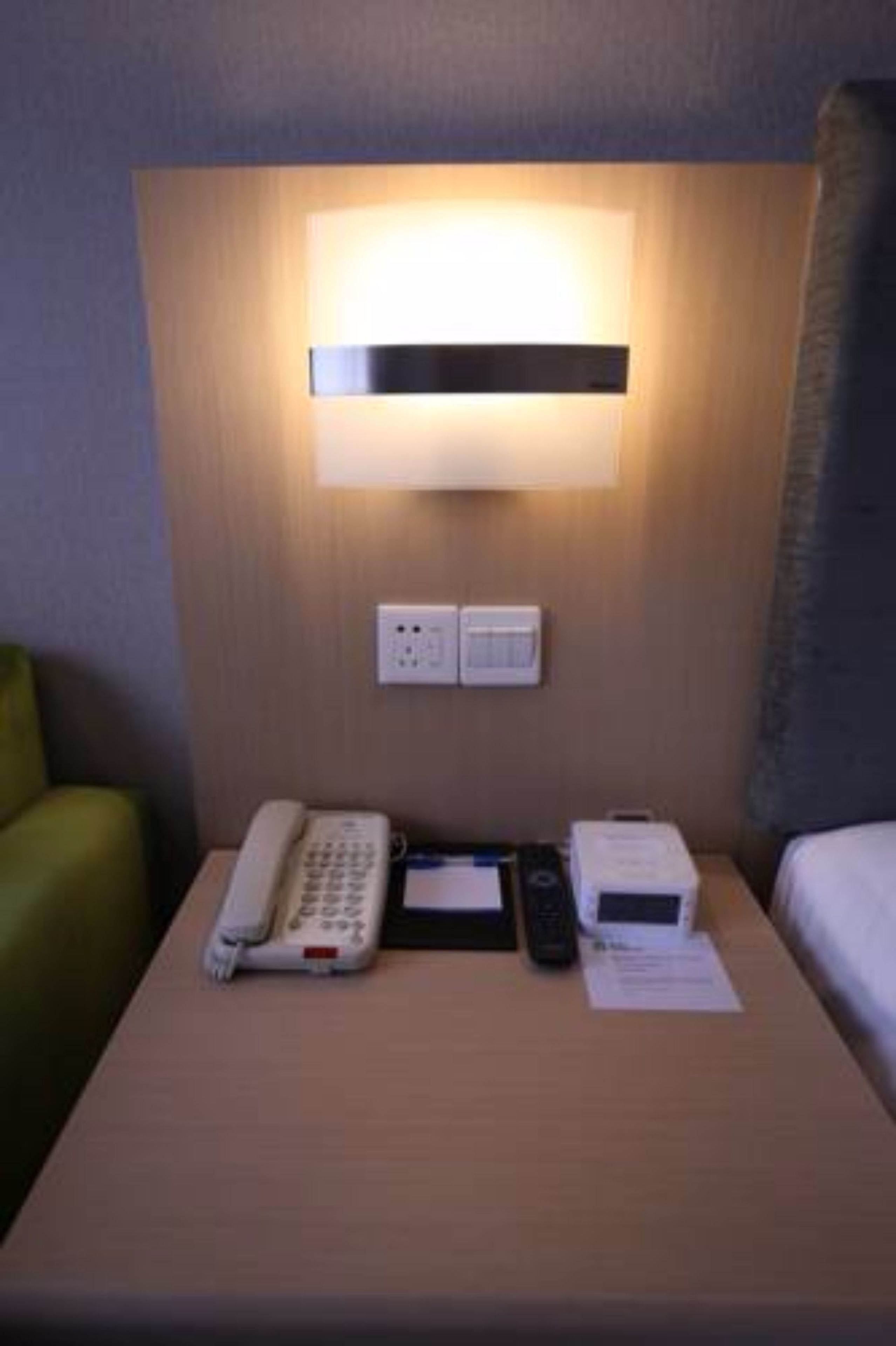 A hotel room with a bedside table featuring a telephone, a remote control, and a small box, possibly a thermostat or control panel.