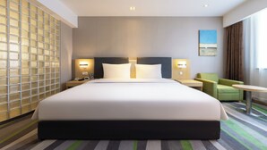 Une chambre d’hôtel avec un grand lit, deux tables de chevet, un fauteuil vert, une petite table ronde et une vue sur l’extérieur.