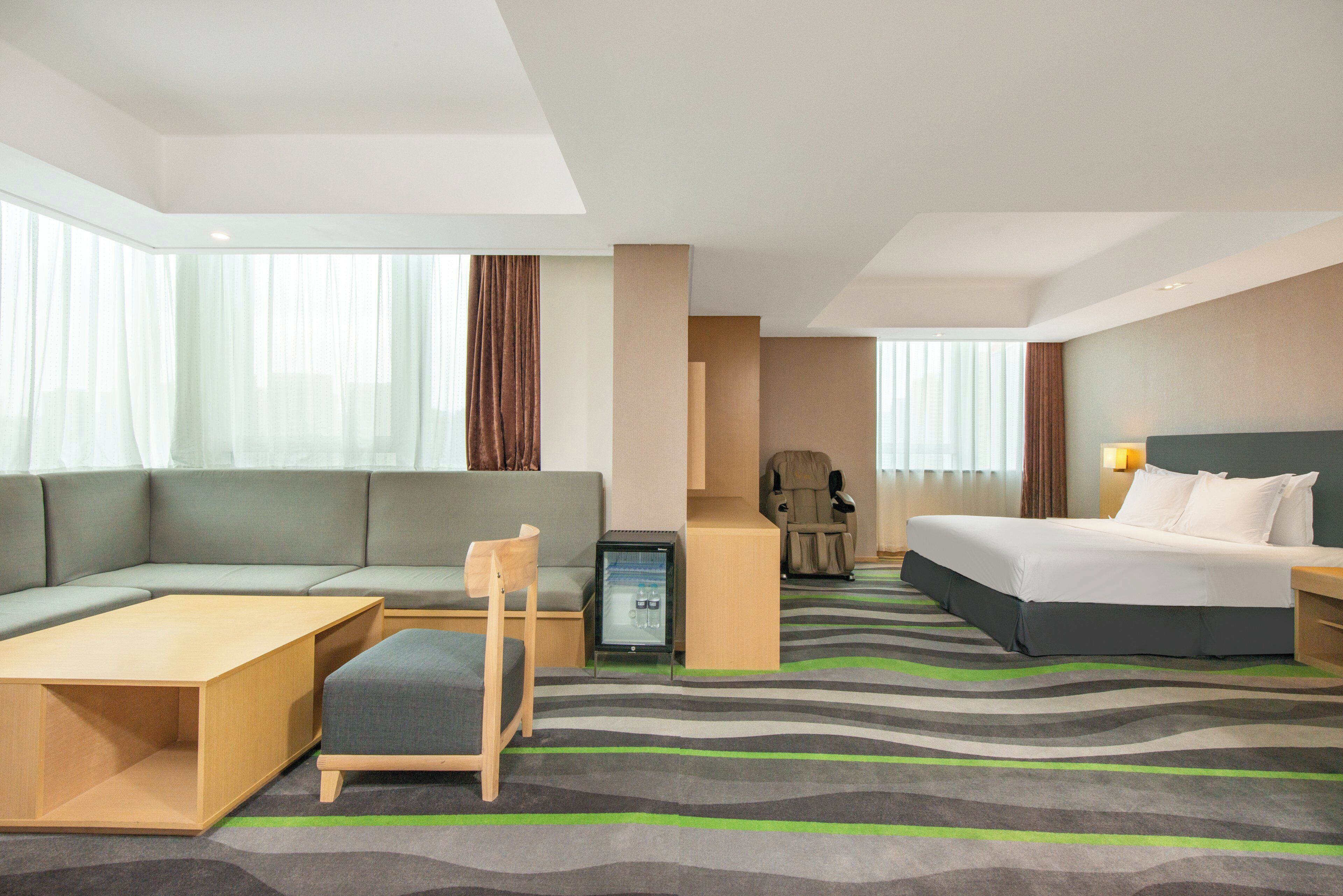 A hotel room with a sofa, a bed, a desk, a chair, a massage chair, and a minibar.