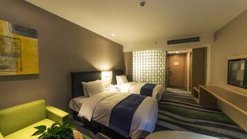 A hotel room with two beds, a seating area with a chair, a television, and a plant.