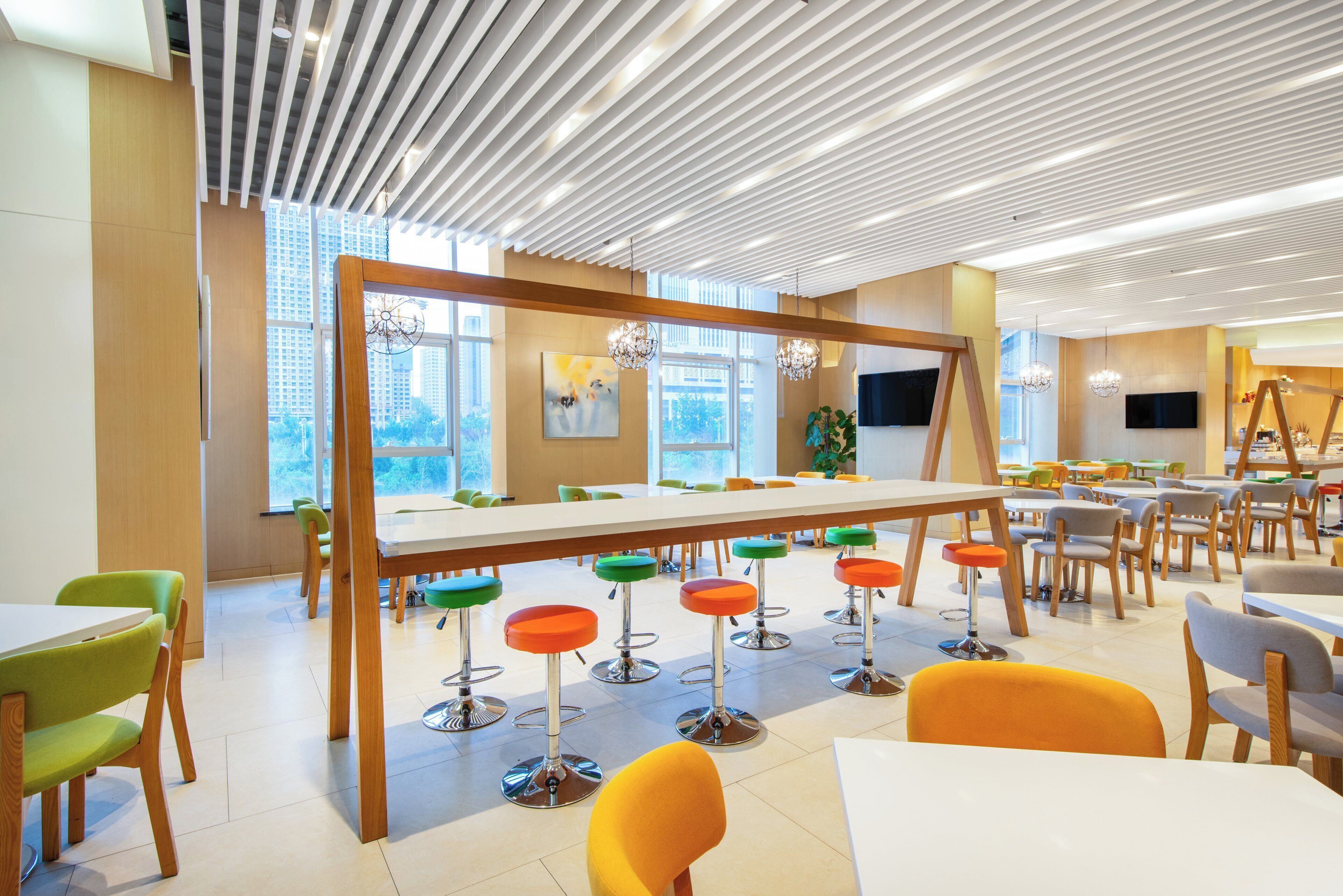 Uma área de refeições moderna com uma mesa comunitária grande, banquetas coloridas e vista para o horizonte urbano através de amplas janelas.