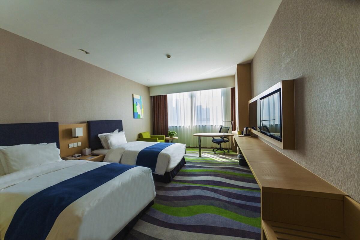 Une chambre d’hôtel avec deux lits, un bureau, une chaise, une télévision et une fenêtre avec des rideaux.