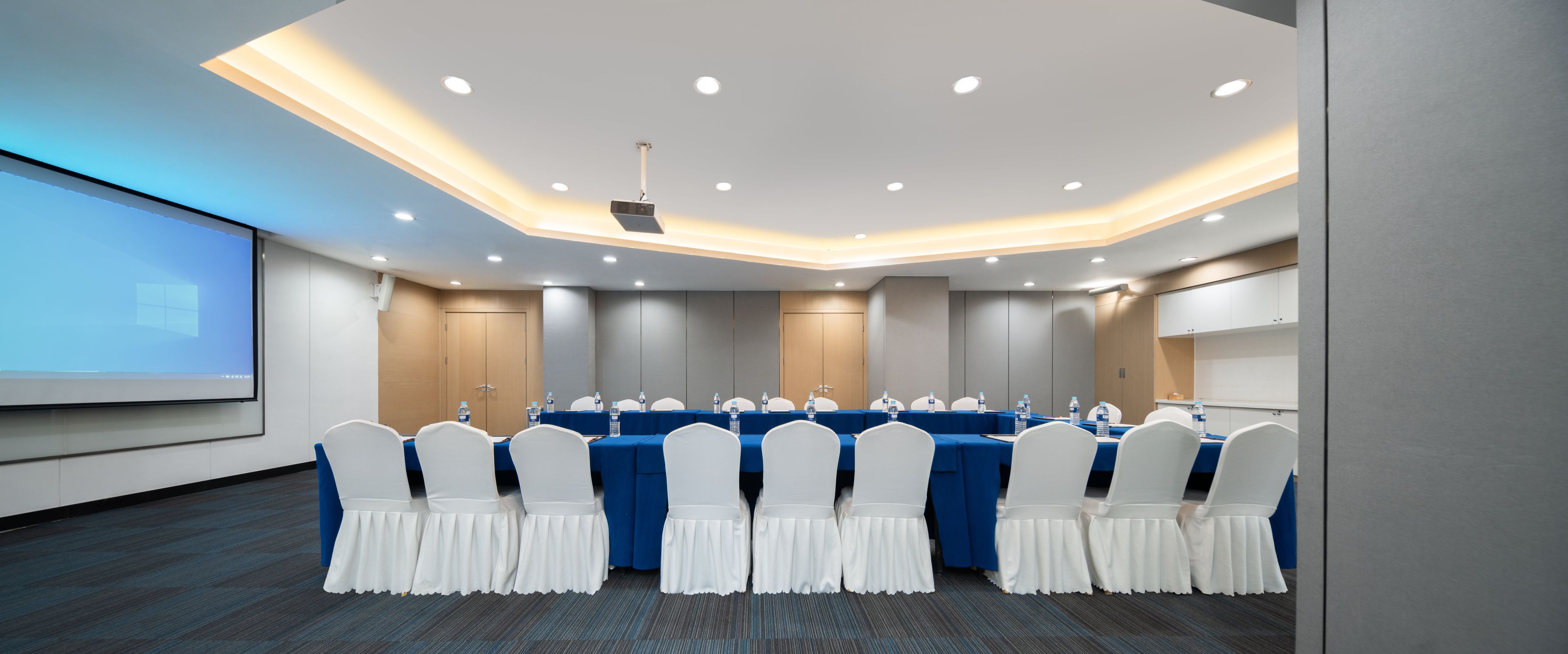 A conference room with a long table, white chairs, and a projector screen.