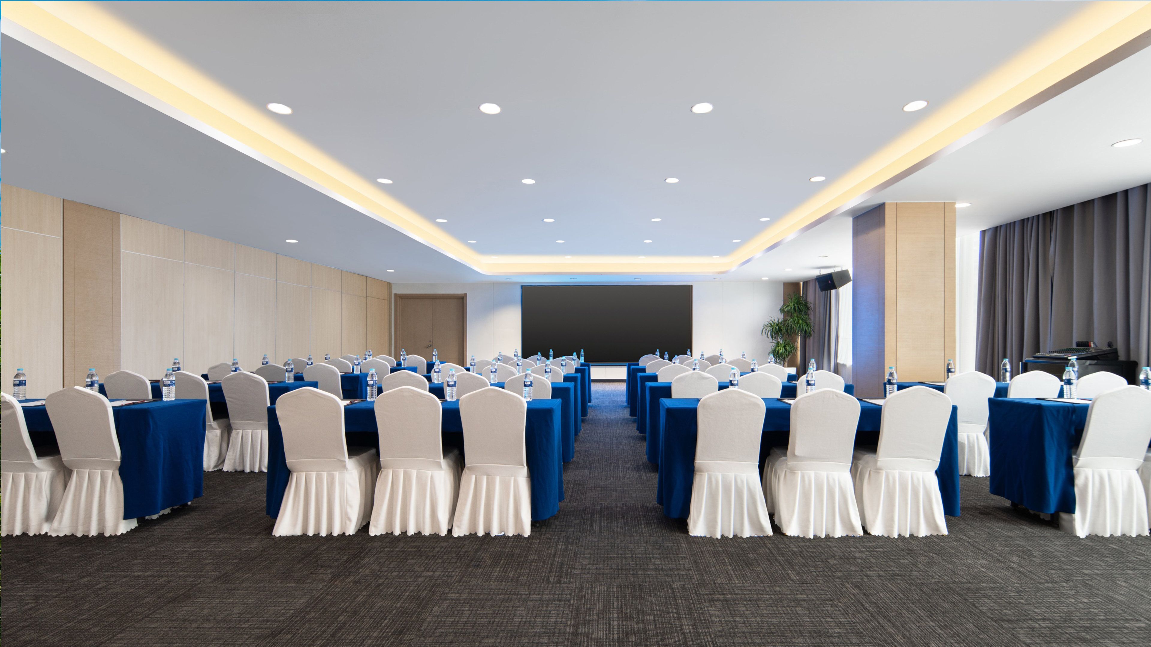 A conference room with rows of tables and chairs, equipped with water bottles, and a large screen at the front.