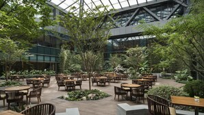 An indoor dining area with multiple tables and chairs, surrounded by greenery and a glass ceiling.