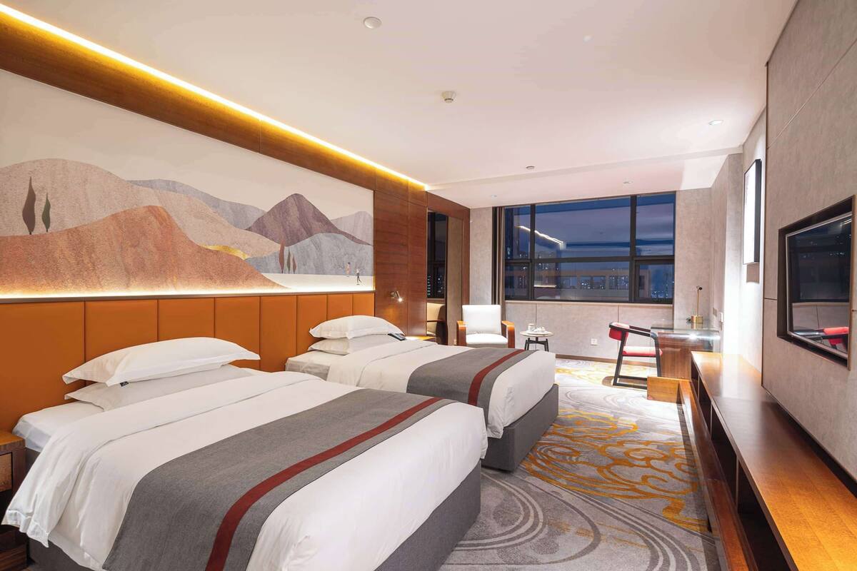 A hotel room with two beds, a desk, a chair, a TV, and a large window with a city view.