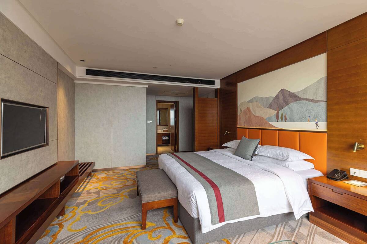 A hotel room with a large bed, a flat-screen TV, a wooden nightstand, a desk, and a painting on the wall.