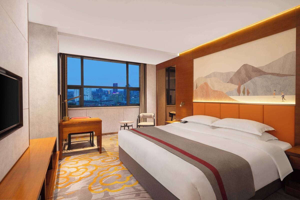 A hotel room with a large bed, a desk, a chair, a TV, and a large window offering a city view.