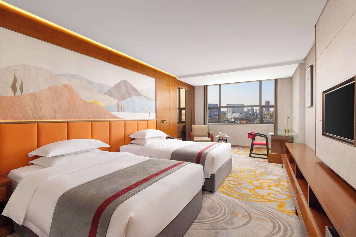A hotel room with two beds, a large window with a city view, a television, and a painting on the wall.