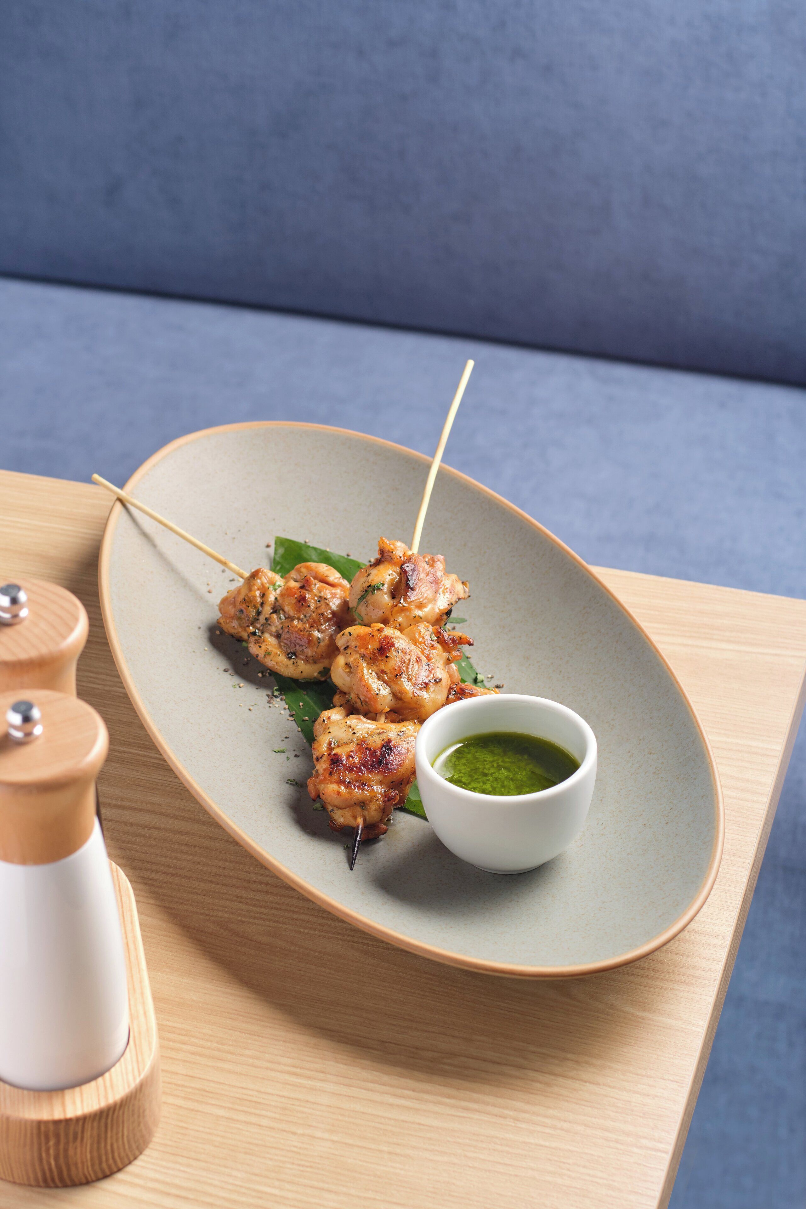 Een bord gegrilde kipspiesjes met een groene saus op een houten tafel.