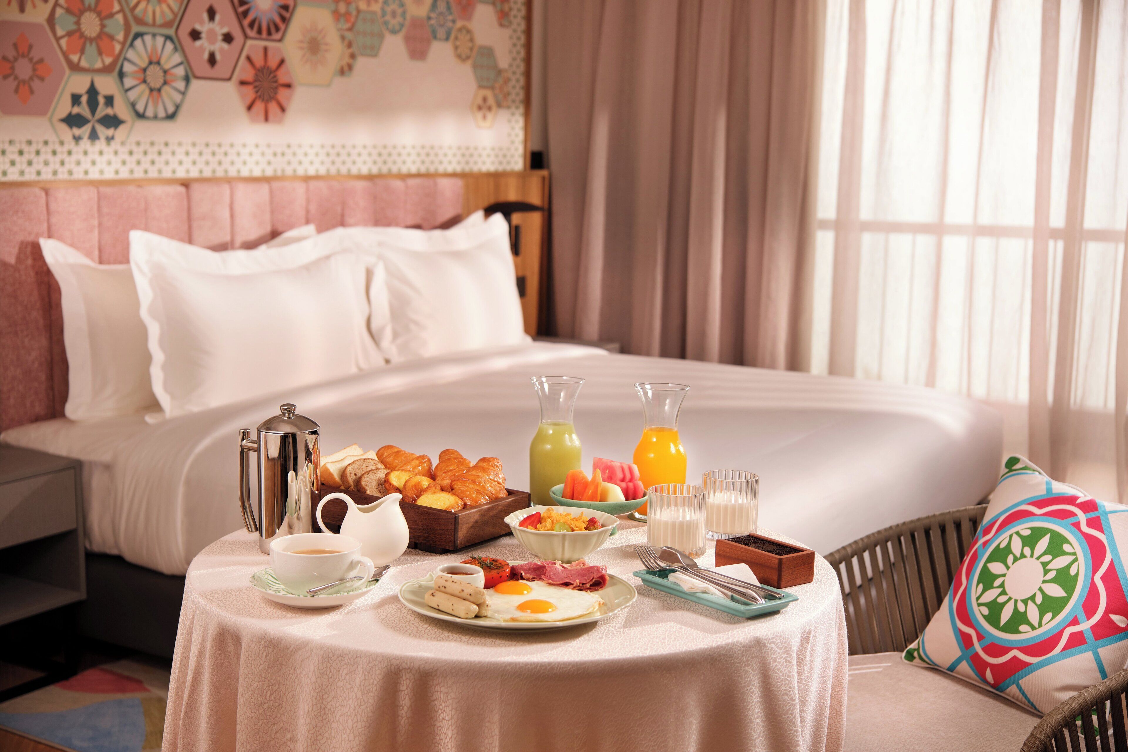 A neatly arranged breakfast table with a variety of food items, a coffee pot, and two juice bottles in a hotel room with a bed and a decorative wall in the background.