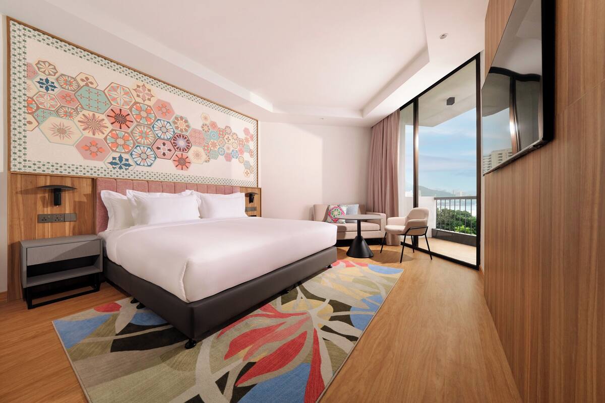 A hotel room with a large bed, a desk, a chair, a TV, and a balcony with a view.