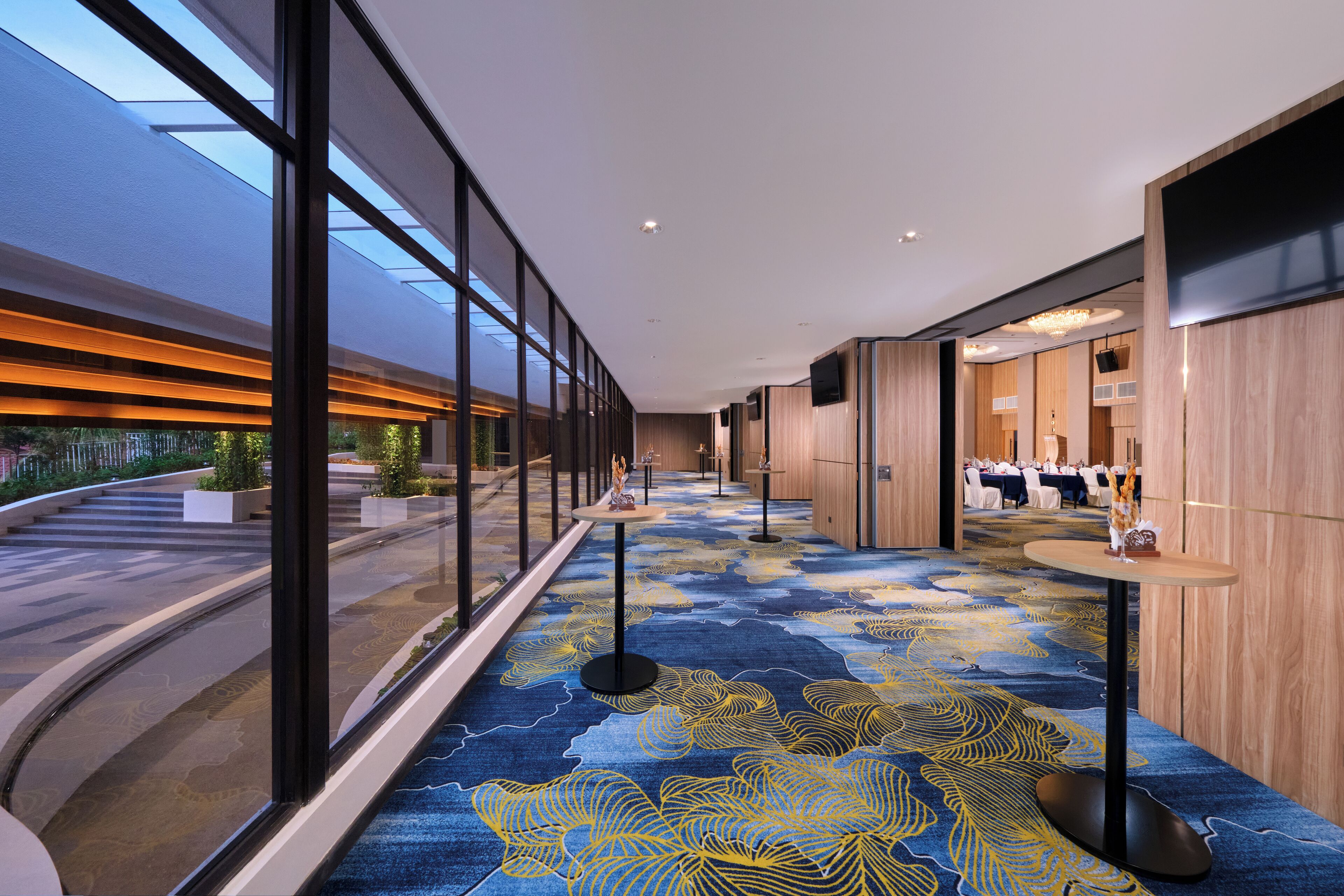 A modern hotel corridor with large windows, a patterned carpet, and a view of the outdoors.