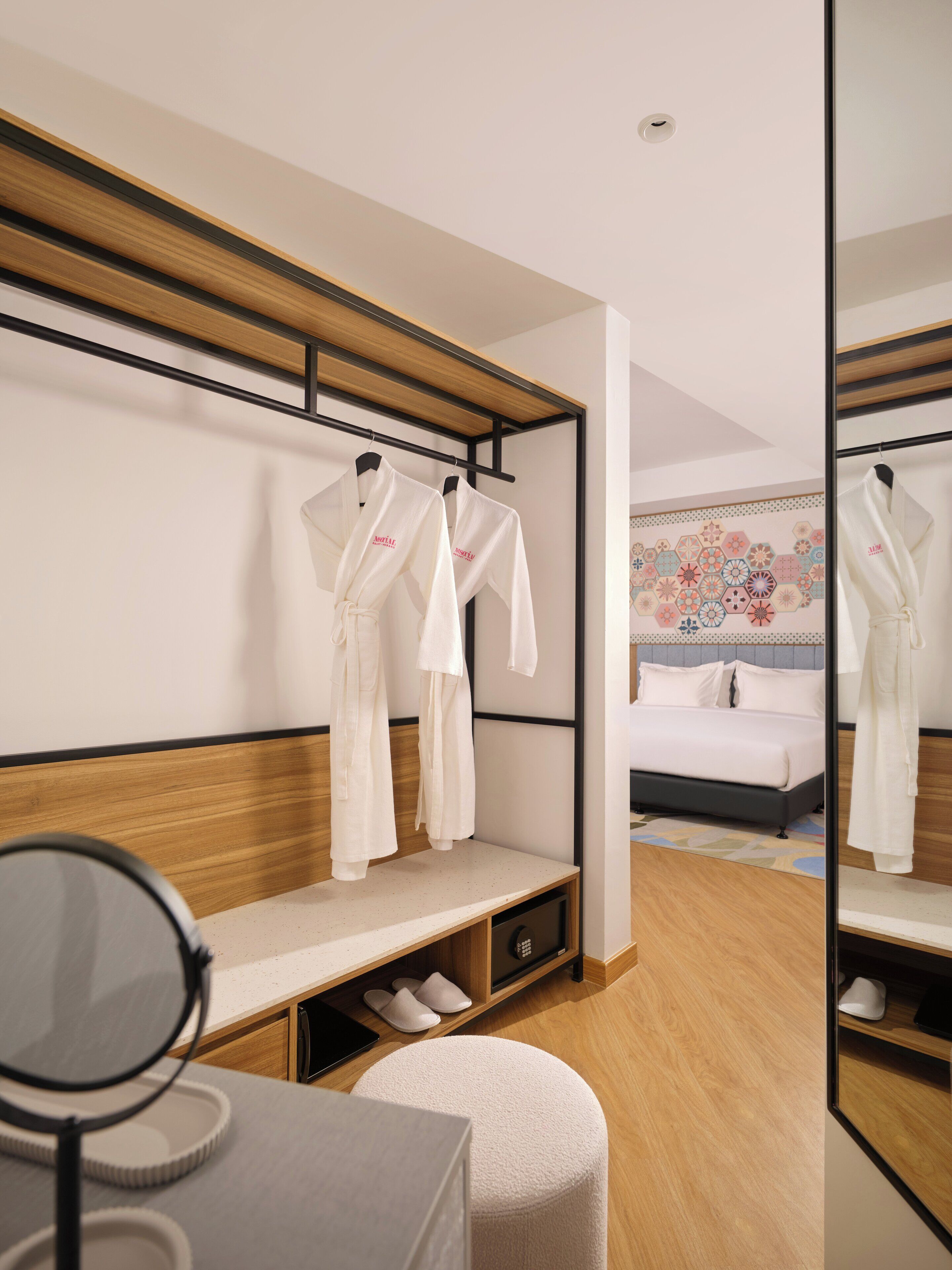 A hotel room with a bed, a bedside table, a mirror, and a bench with robes hanging above.