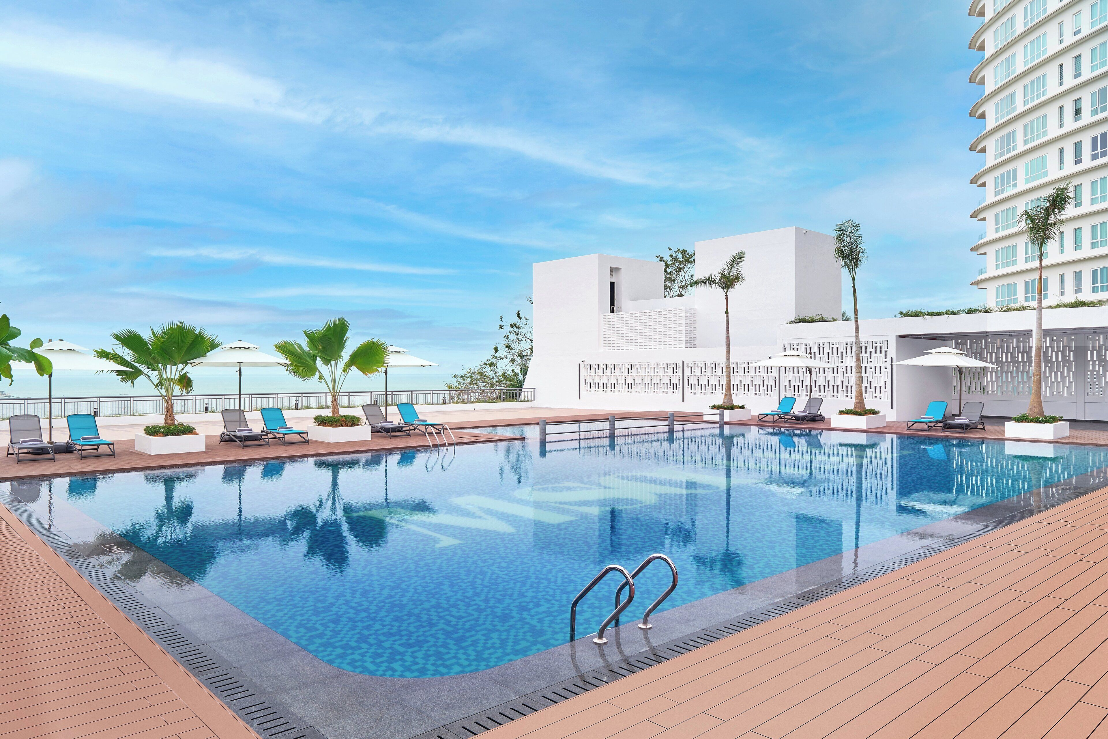 Ein Pool mit Liegestühlen und Palmen, umgeben von einem modernen Gebäude mit Blick auf den Ozean.