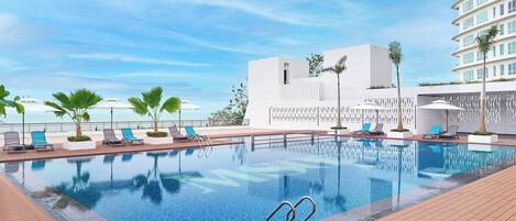 A swimming pool with lounge chairs and palm trees, surrounded by a modern building with a view of the ocean.