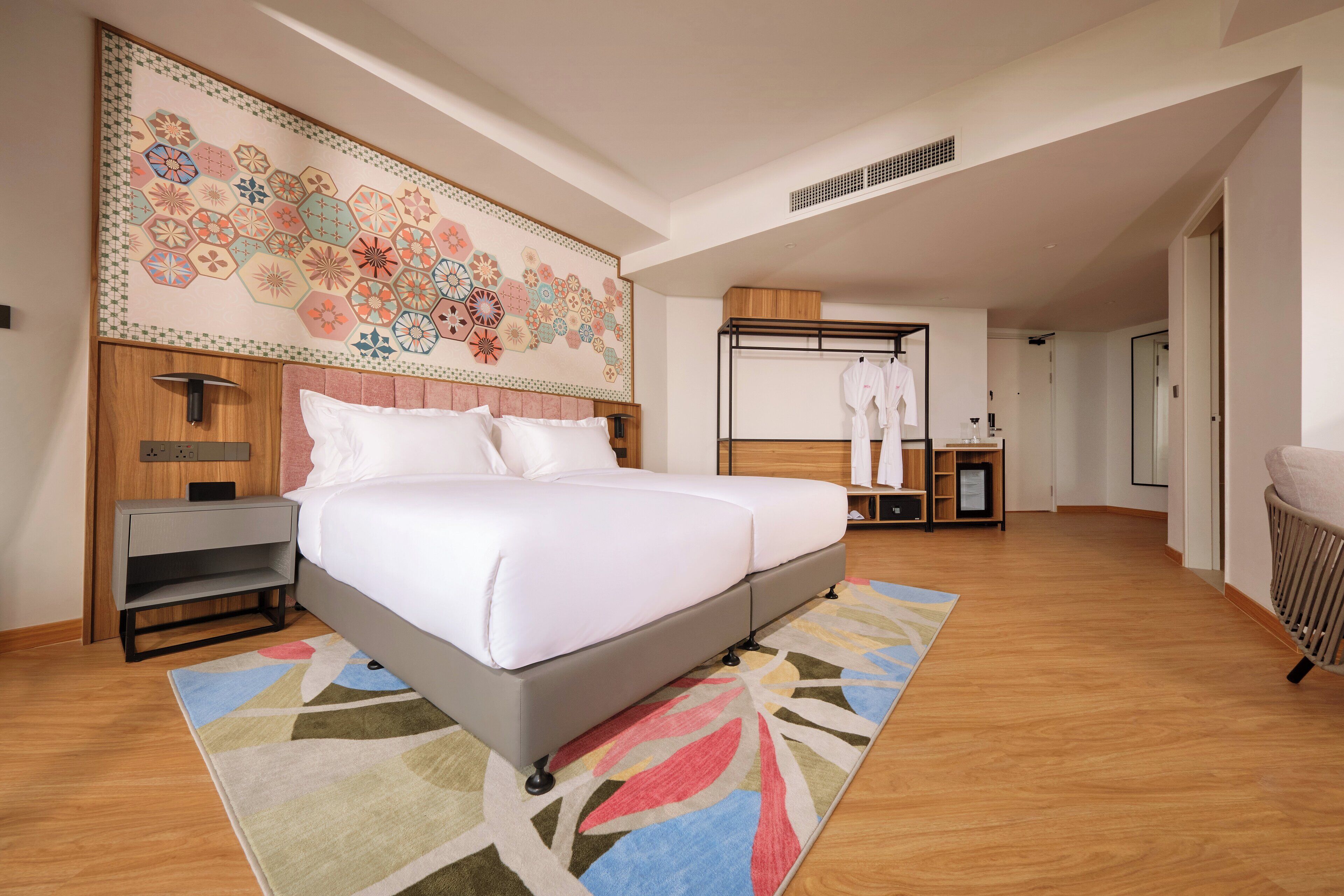Une chambre d’hôtel moderne avec un grand lit, une table de chevet, une tête de lit en bois, un tapis coloré et une œuvre d’art murale.
