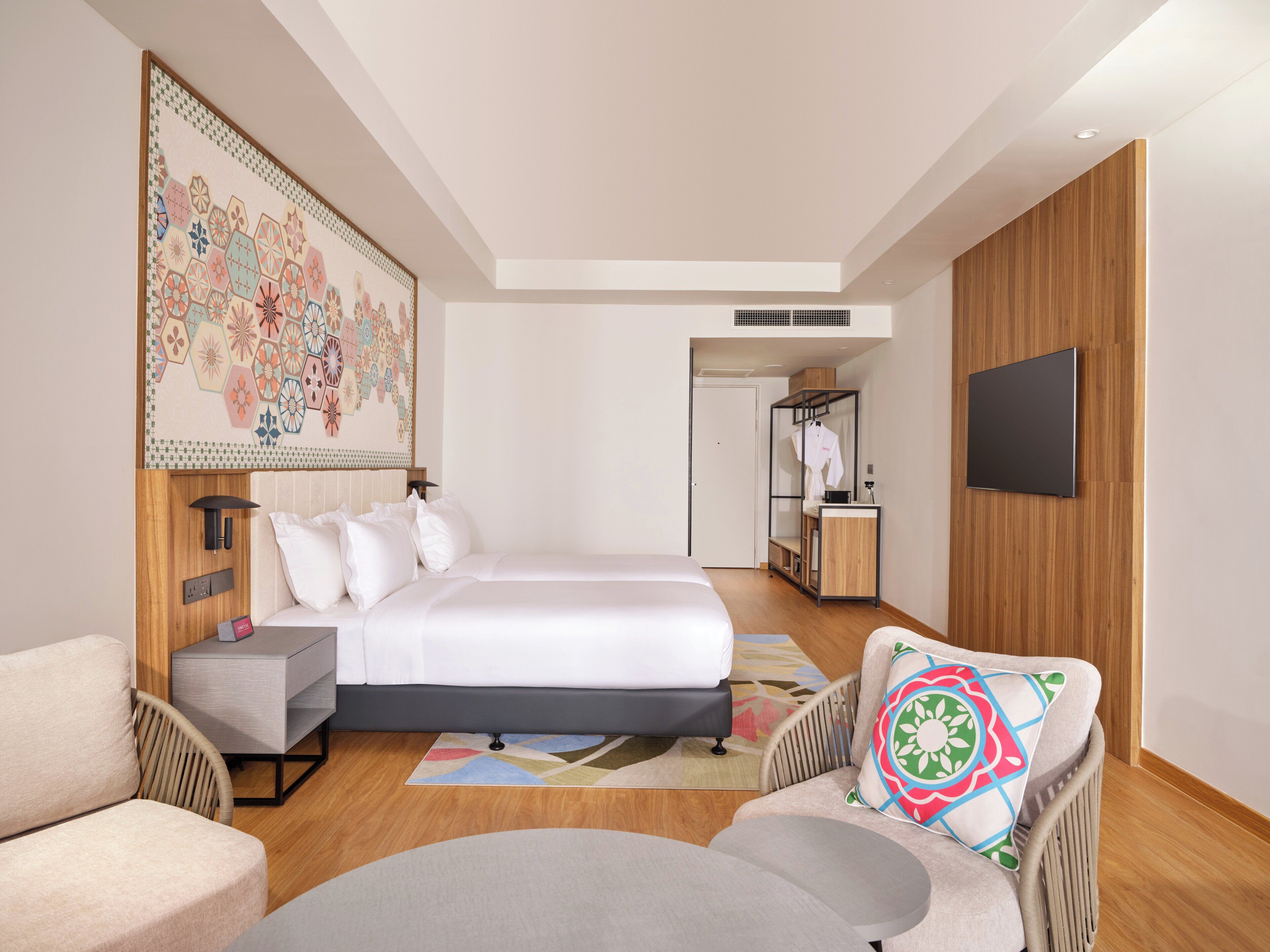 A modern hotel room with a large bed, a seating area with a chair and sofa, a small table, a TV mounted on the wall, and a wooden panel design on the walls.
