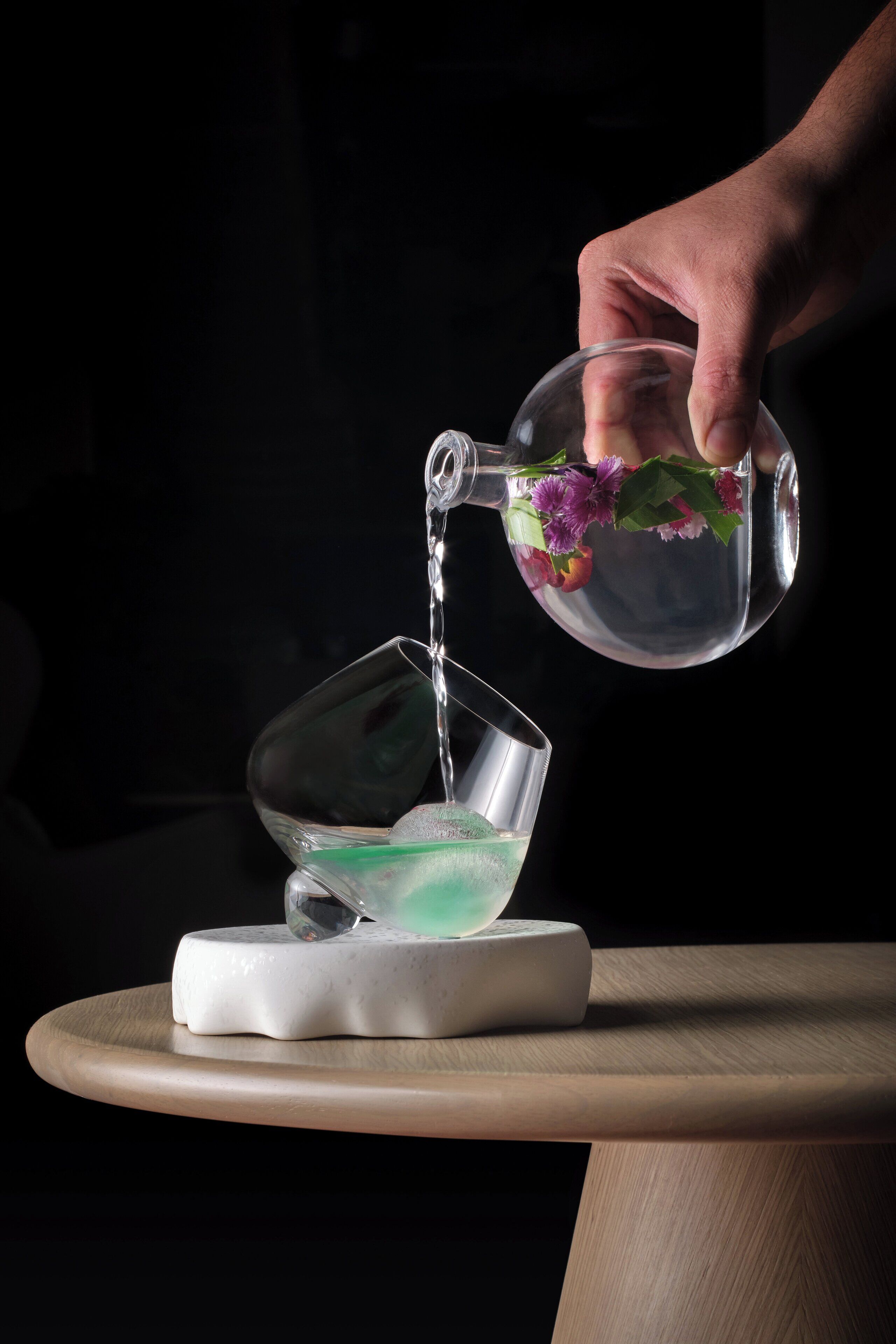 A hand pouring water from a glass pitcher into a glass on a white coaster.