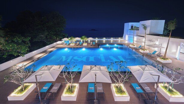 A swimming pool with lounge chairs and umbrellas, surrounded by trees and a view of the sea. - M Social Resort Penang (George Town)