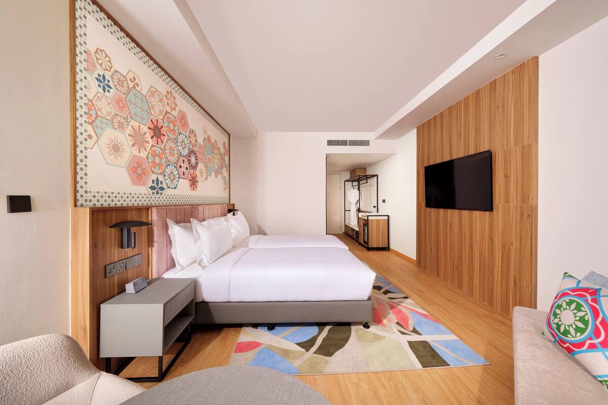 A modern hotel room with a large bed, a desk, a flat-screen TV mounted on the wall, and a colorful rug.
