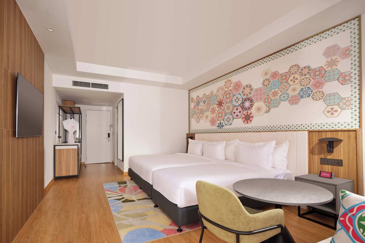 A modern hotel room with a large bed, a round dining table, a chair, a TV, and a bathroom visible through an open door.