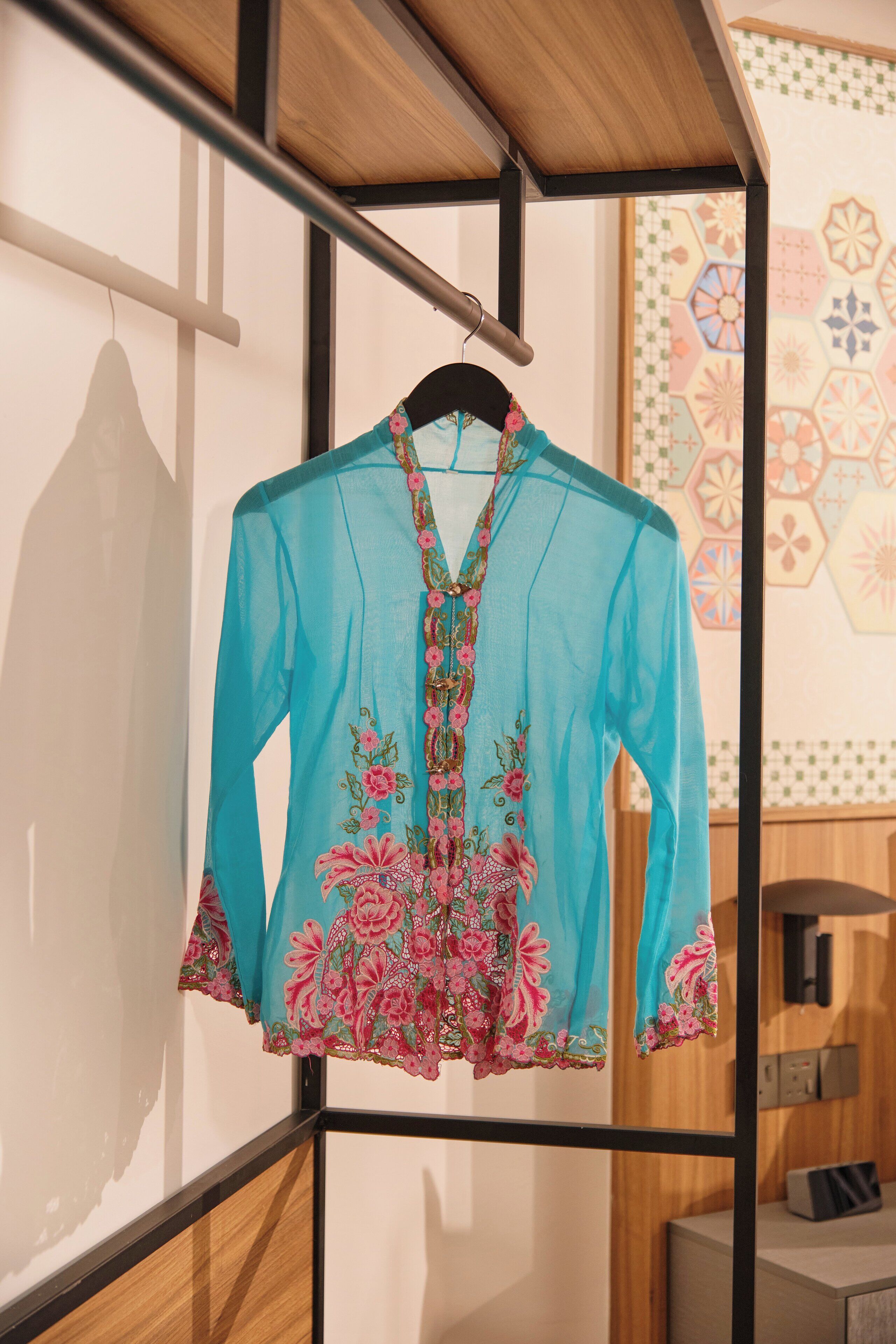 Eine blaue Bluse mit Blumenstickerei, die an einem Kleiderbügel in einem Raum mit Holzvertäfelung und Wandverkleidung mit geometrischen Mustern hängt.