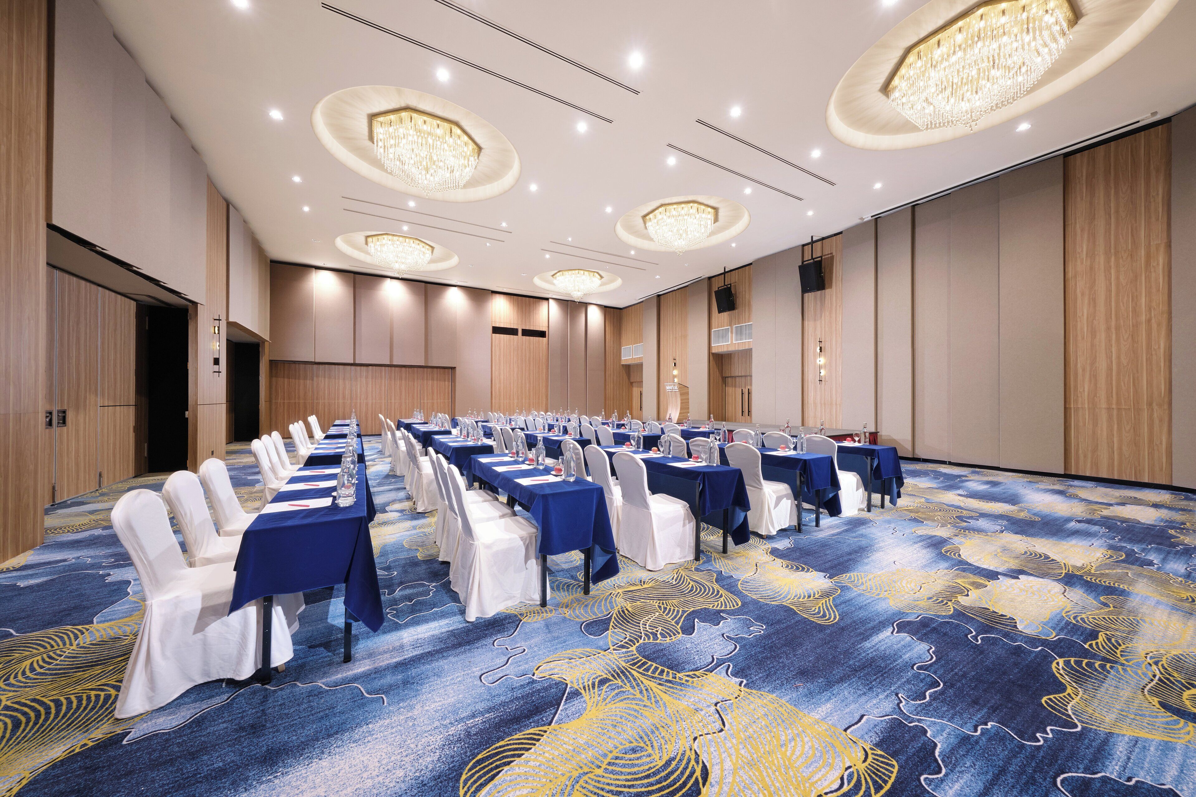 Un salón de conferencias con filas de mesas y sillas, una alfombra estampada y candelabros en el techo.