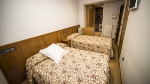 Habitación de hotel con dos camas, cabecero de madera, una mesita de noche pequeña, un televisor instalado en la pared y una puerta que da a un baño.