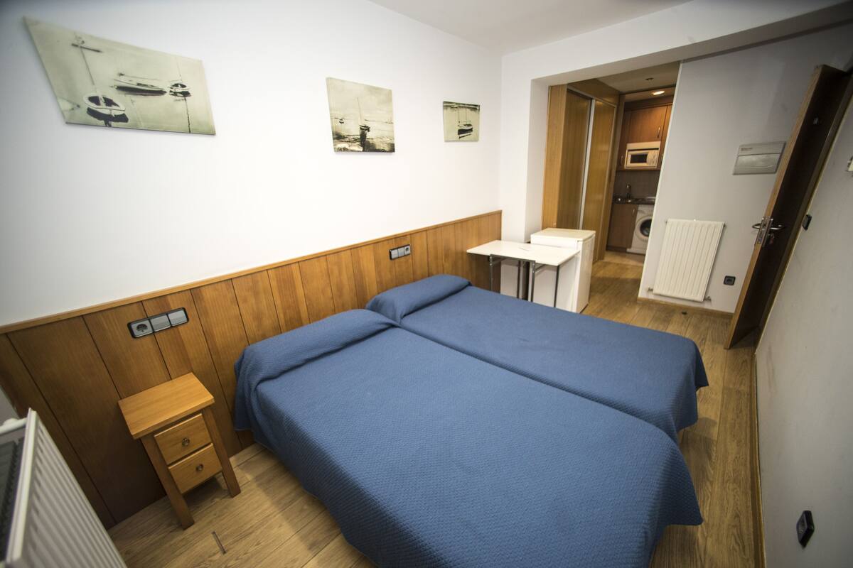 Quarto com duas camas, uma mesa de cabeceira de madeira, uma mesinha e vista para uma cozinha e um banheiro através de uma porta aberta.