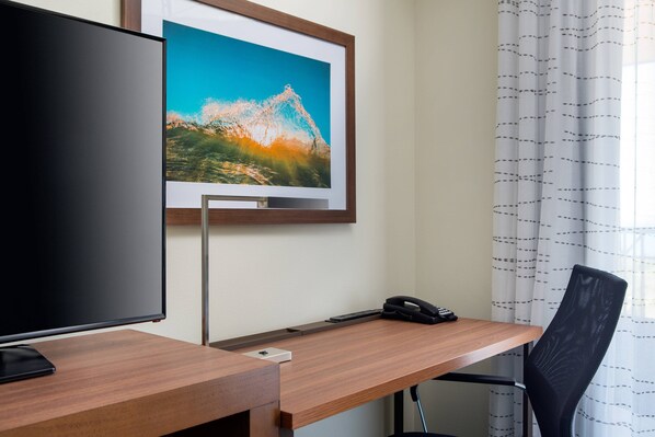 A hotel room with a wooden desk, a framed picture of a mountain, a black chair, and a telephone.