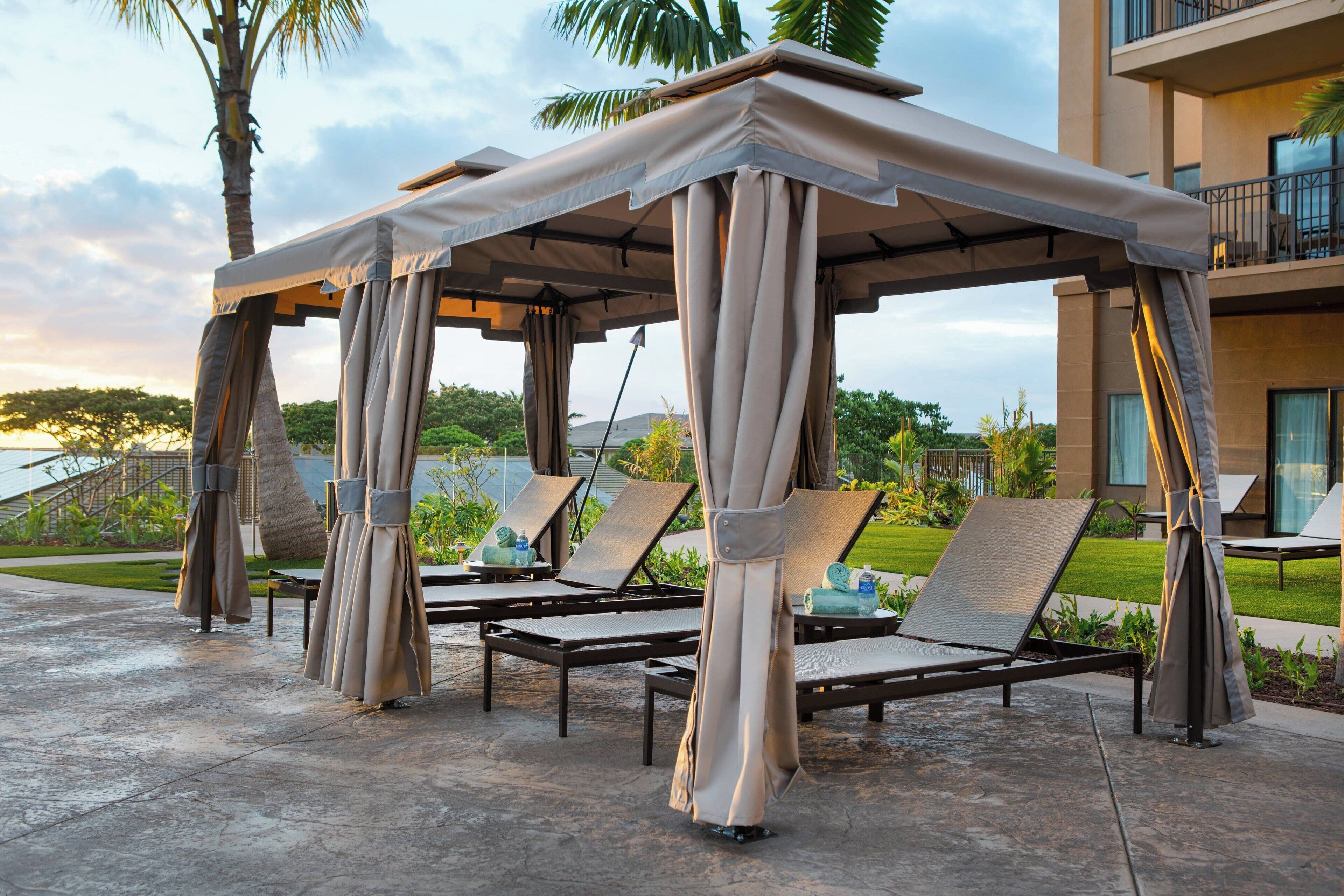 A covered outdoor seating area with lounge chairs and umbrellas, set against a backdrop of a modern building and palm trees.