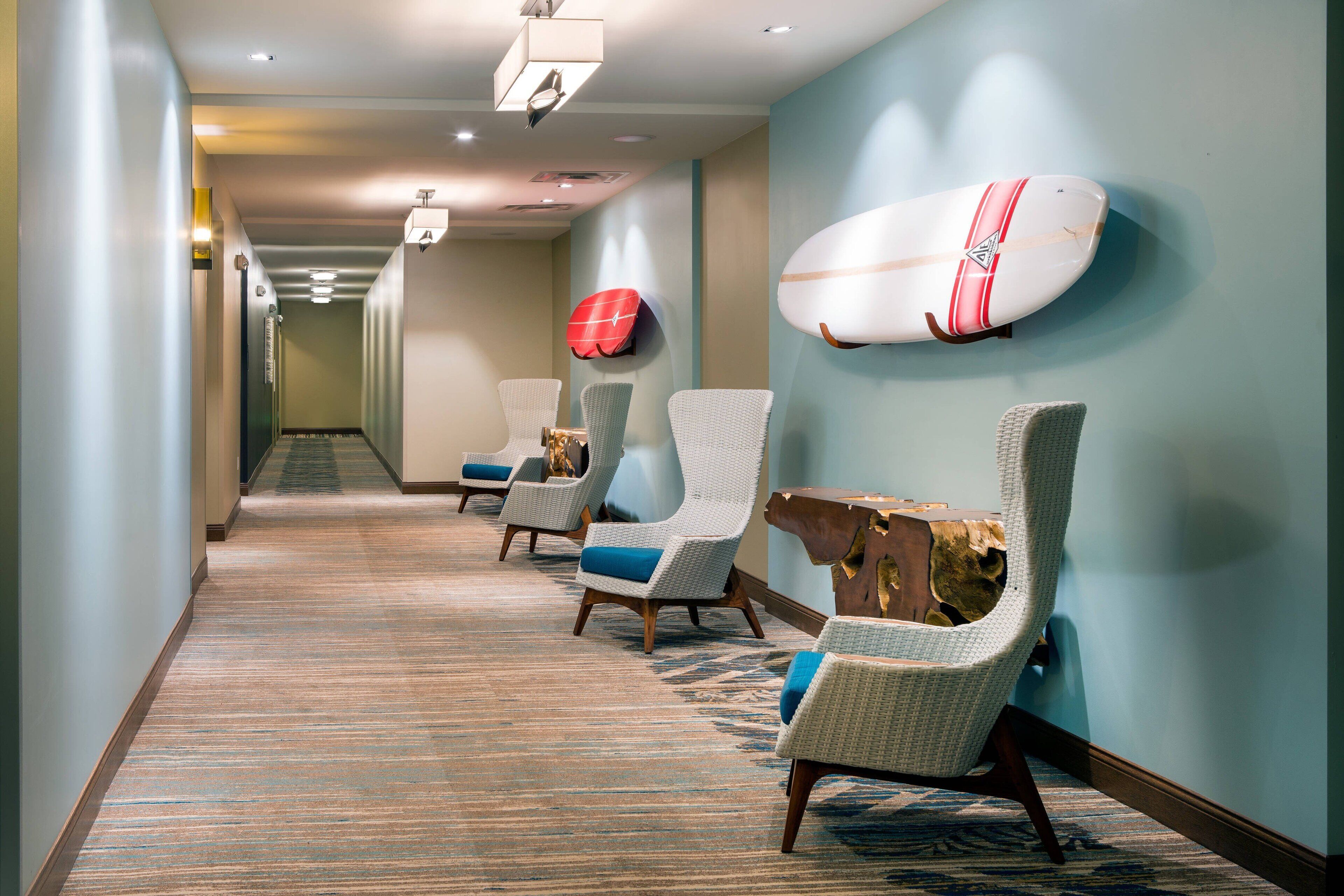 A hotel hallway with a carpeted floor, three wicker chairs with blue cushions, and a surfboard mounted on the wall.
