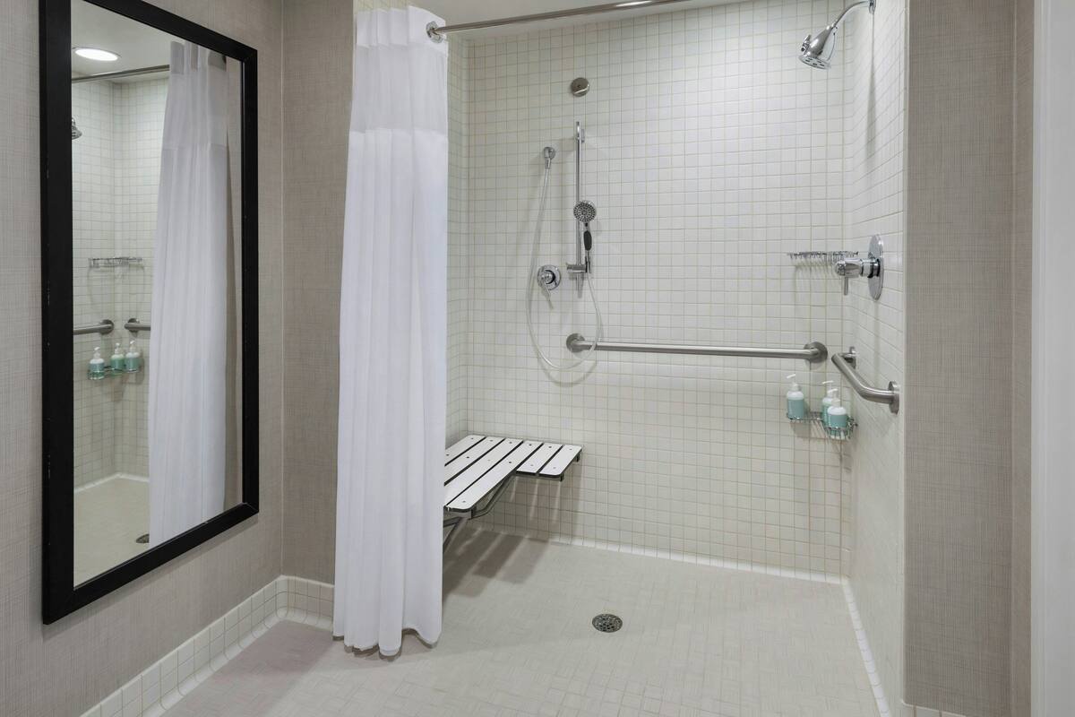 A walk-in shower with a white tiled floor, a shower control panel, a handheld showerhead, a handheld shower attachment, a grab bar, and a bench.