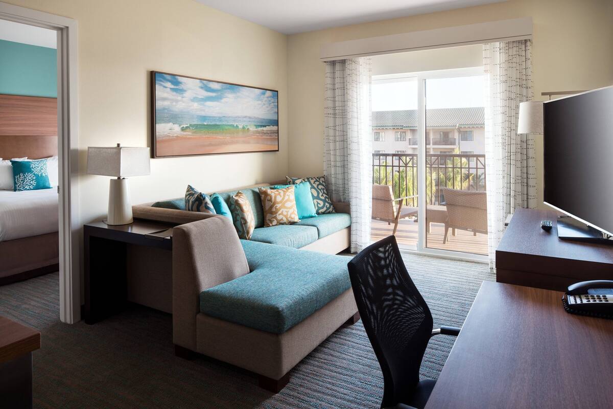 A hotel room with a bed, a sofa, a desk, a chair, and a balcony with a view of the ocean.