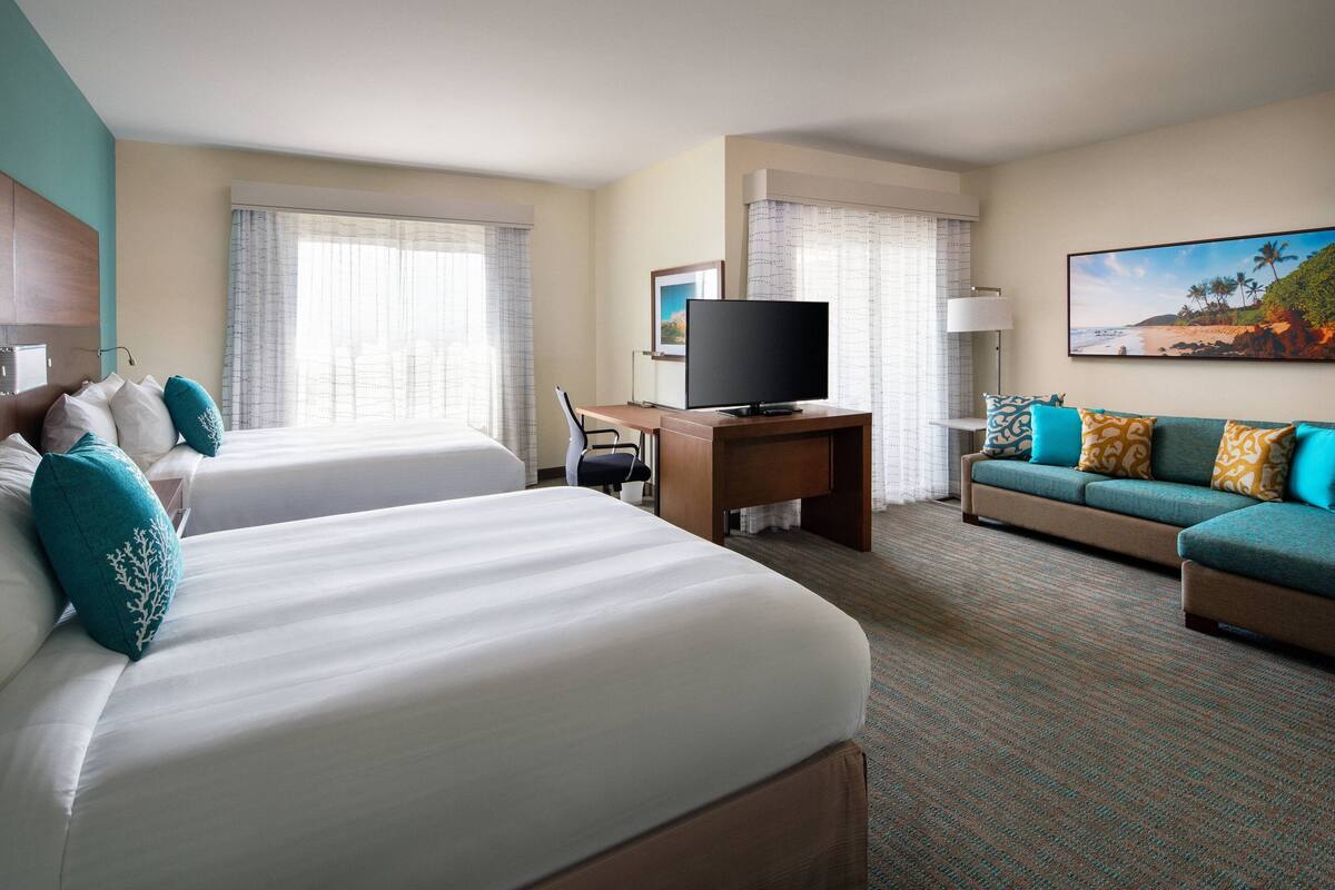 A hotel room with two beds, a desk with a TV, a sofa, and a large window with curtains.